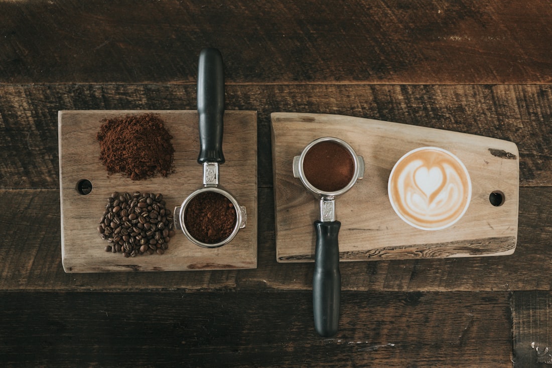 Freshly ground coffee, whole beans and a portafilter laid out on a dark wooden bench beside a finished cappuccino.