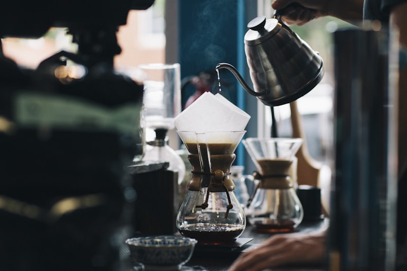 A barista pouring from a gooseneck kettle into a pour-over brewer, steam rising off the bed of coffee.