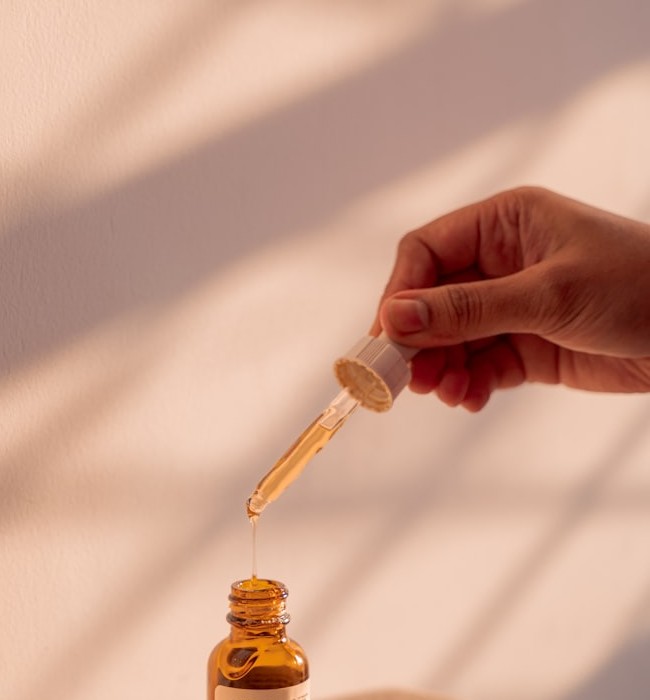 A dropper of skincare serum held above an amber bottle, warm afternoon light